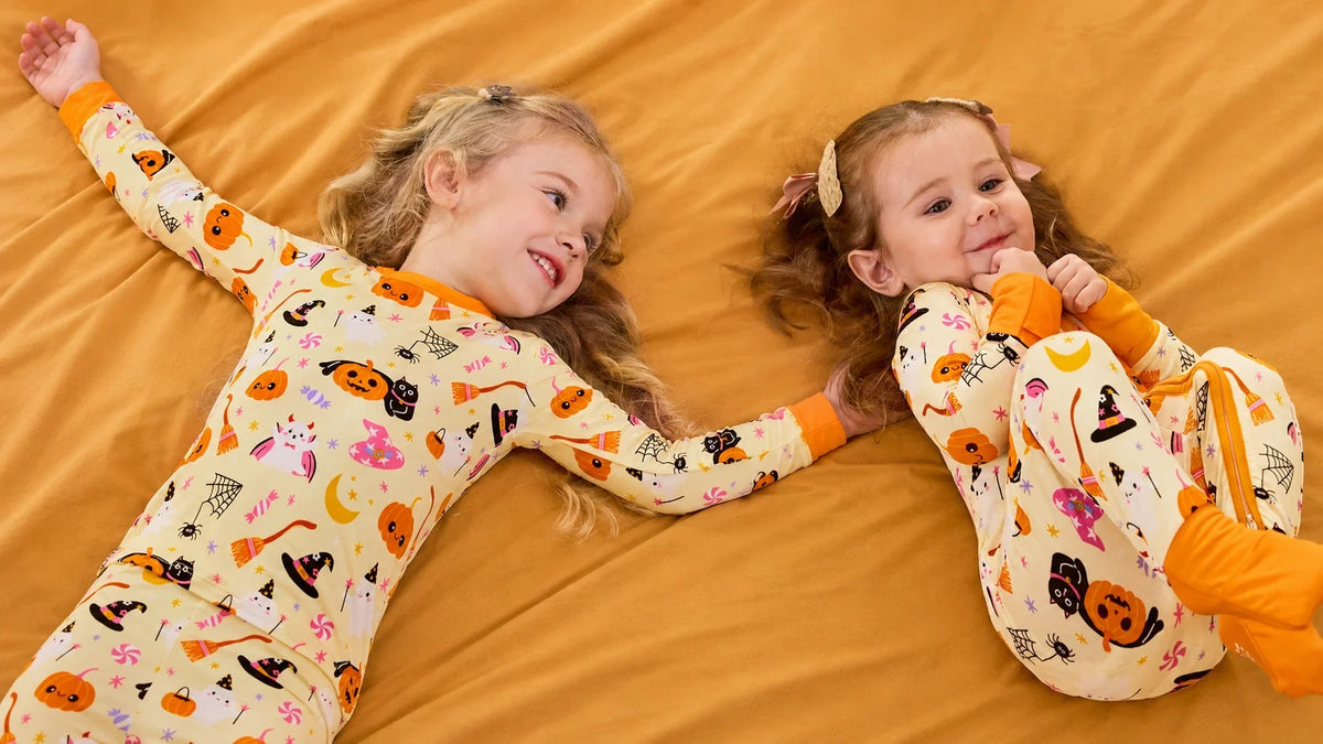 Two siblings holding hands in coordinating pumpkin-print Halloween pajamas on a golden blanket