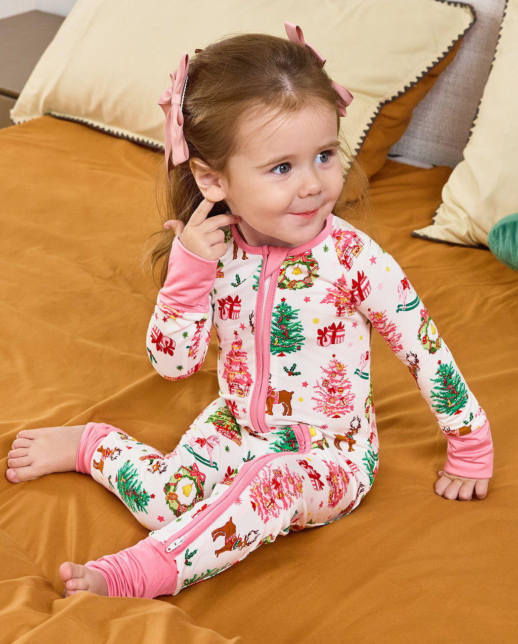 Child seated on bedding with a hand to the cheek in the Blush Christmas romper.