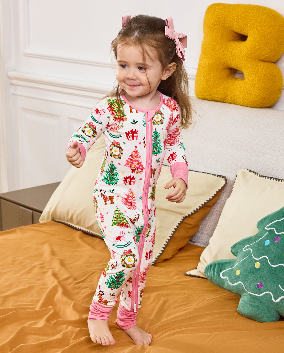 Child standing on bedding with a hand raised in the Blush Christmas romper.