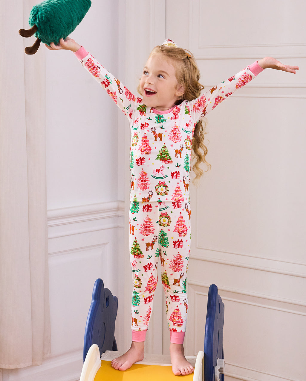 Child standing with arms raised and a plush Christmas tree in Blush Christmas pajamas.