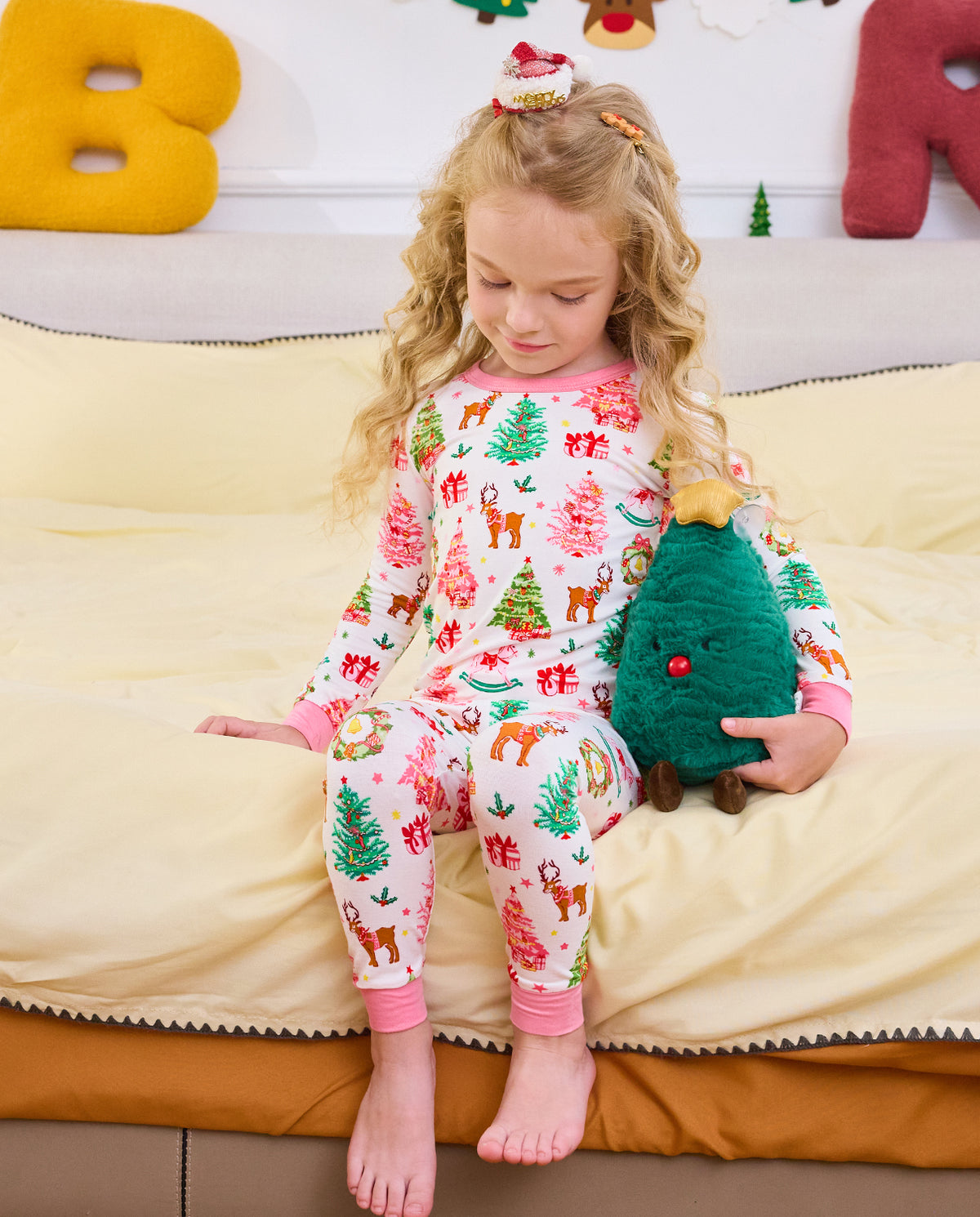 Child looking down and holding a plush tree while seated on a sofa in Blush Christmas pajamas.
