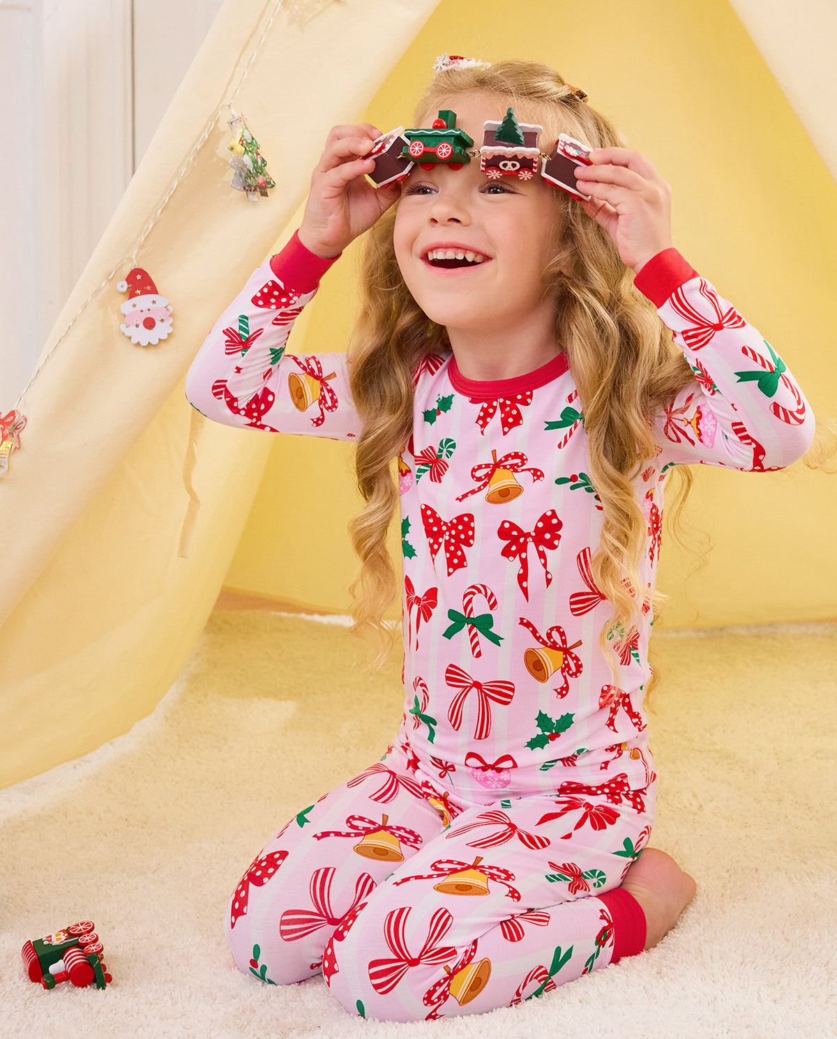 Child seated by a play tent with a toy train held above the eyes in Christmas Bow pajamas.
