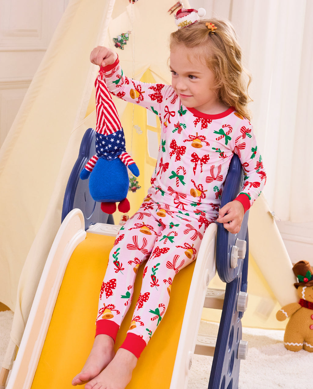 Child seated on a slide holding a plush toy in Christmas Bow pajamas.
