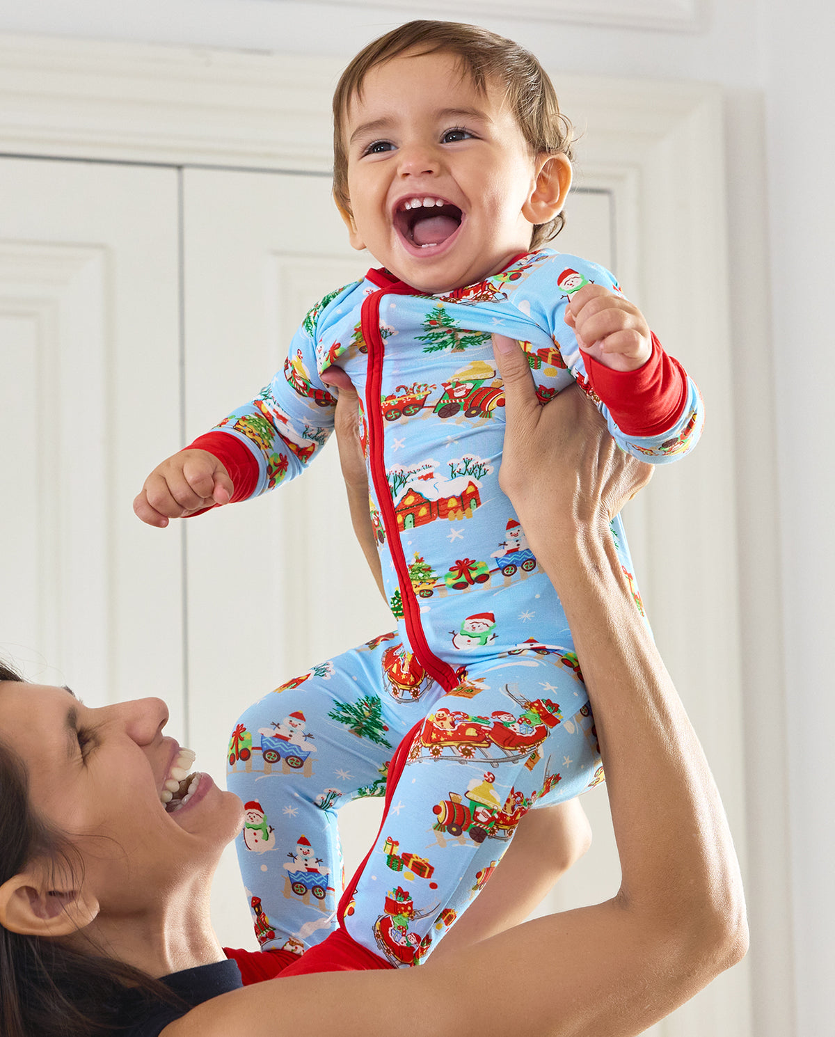 Parent lifting a smiling child wearing the Christmas Trains Bamboo Zippy with festive train print and red trim.