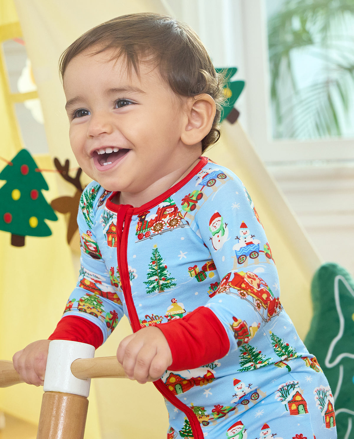 Smiling child wearing the Christmas Trains Bamboo Zippy while holding the handlebars of a wooden ride-on toy.