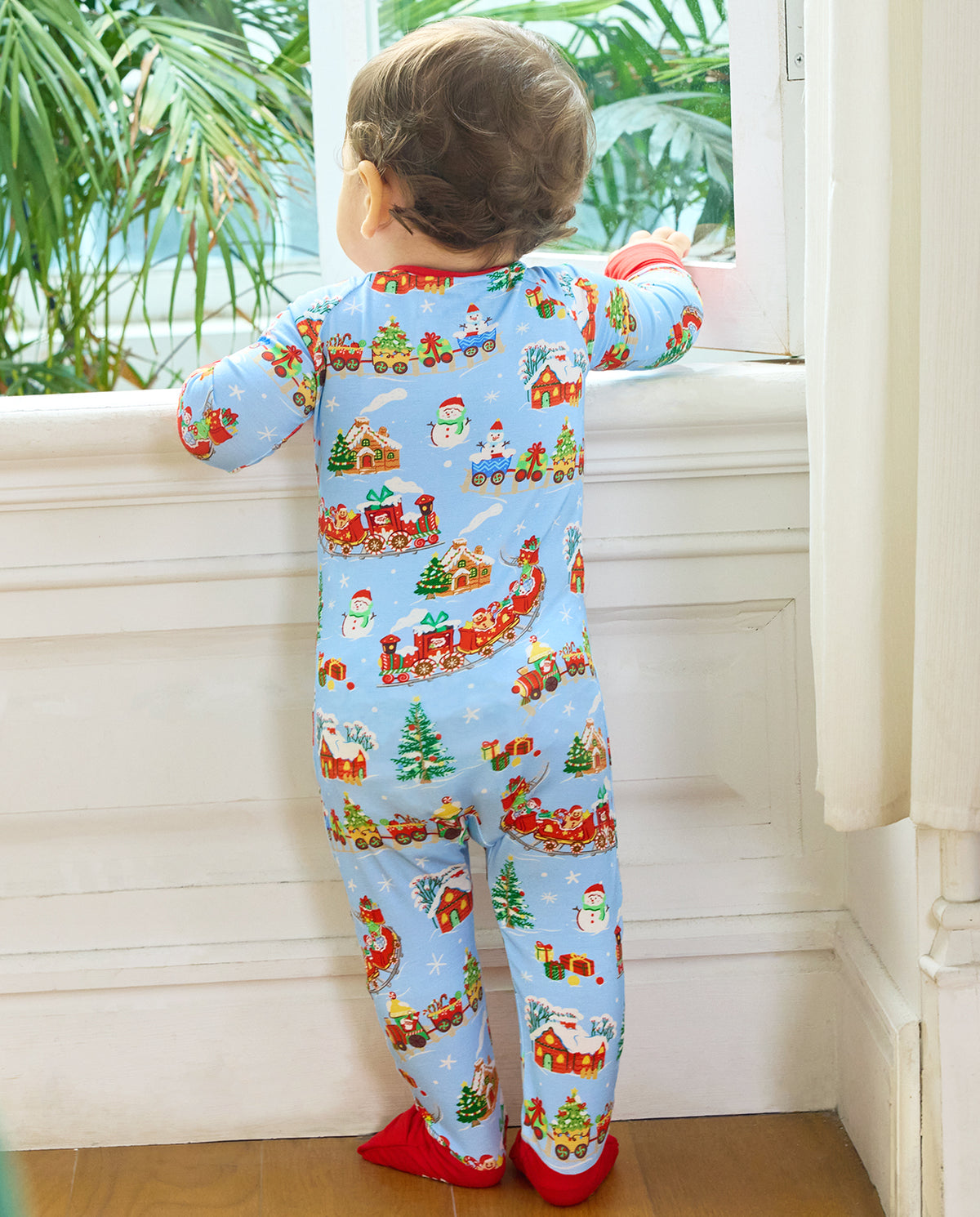 Full back view of a child standing at a window in the light blue Christmas Trains Bamboo Zippy with red foot cuffs.