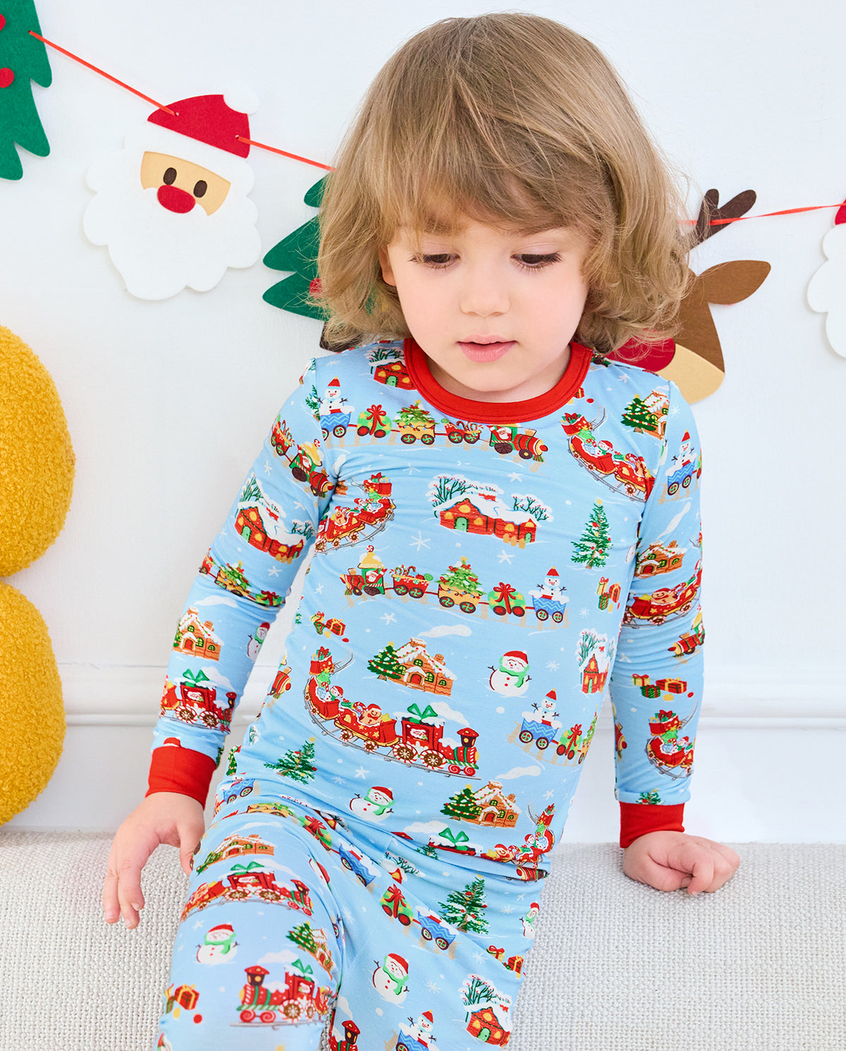 Child seated in light blue Christmas Trains pajamas with red cuffs.