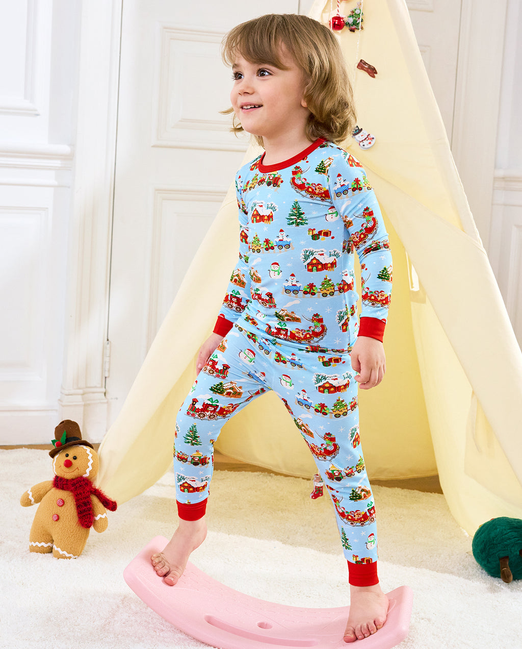 Child standing by a pink rocker beside a holiday play tent in Christmas Trains pajamas.