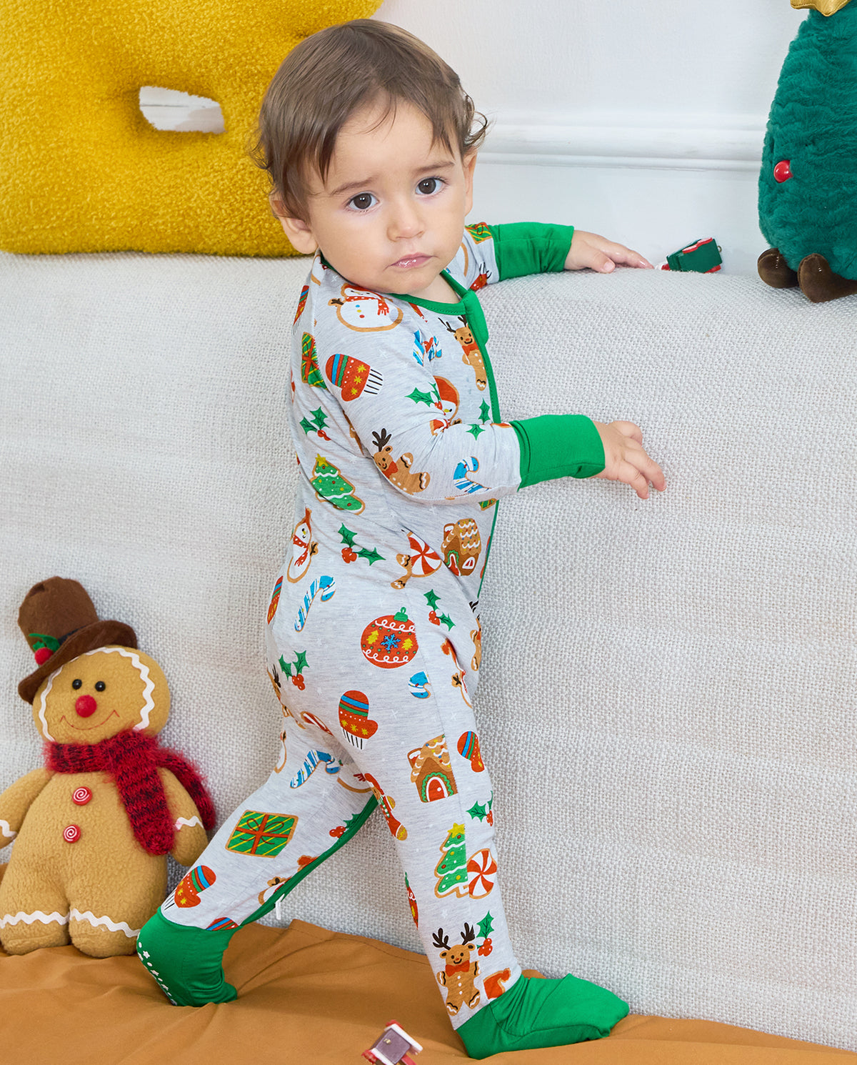 Baby standing beside a sofa in the Gingerbread Gala Bamboo Zippy, showing the Christmas print and covered green feet