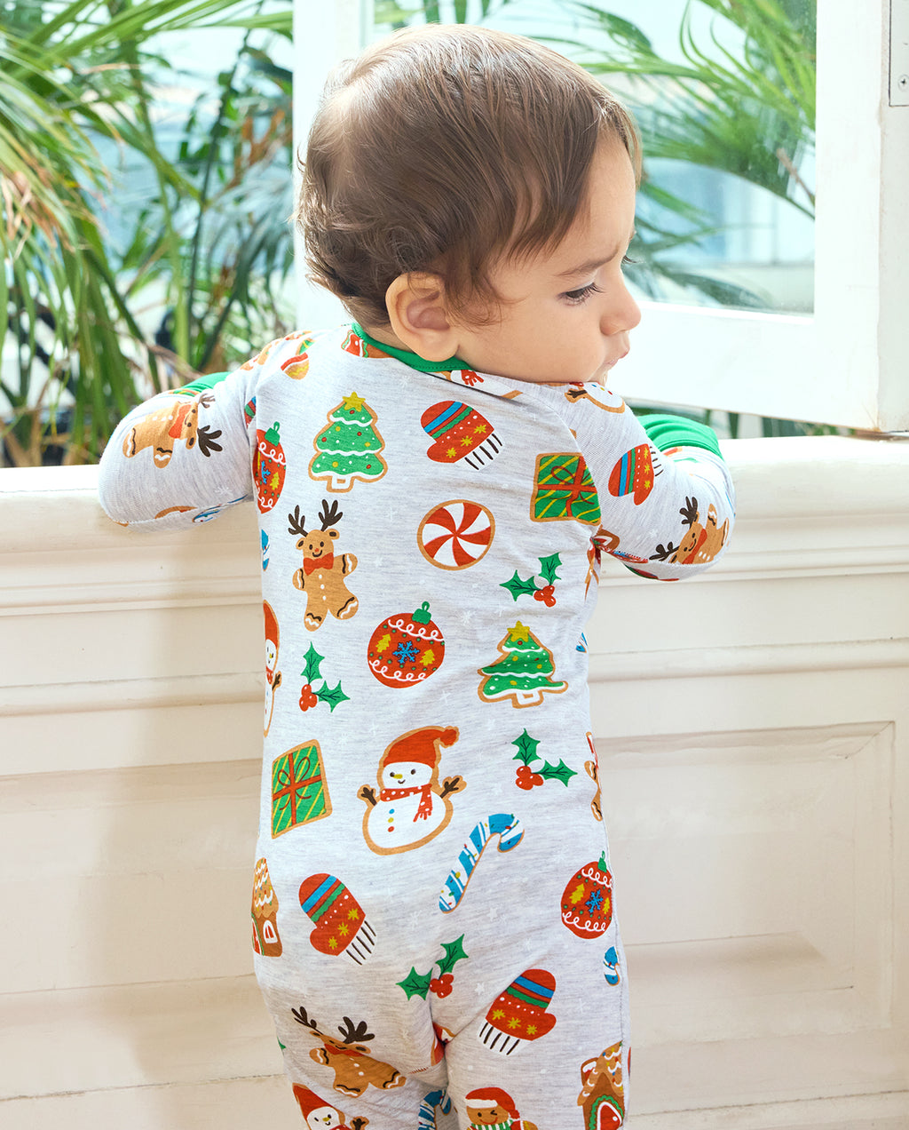 Back view of a baby wearing the Gingerbread Gala Bamboo Zippy by a window, showing gingerbread reindeer and Christmas trees