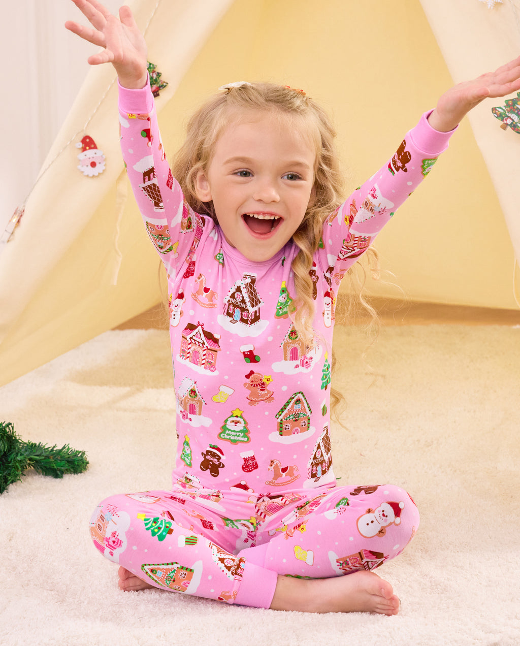 Child sitting on bedding with arms raised in Gingerbread Lane pajamas.
