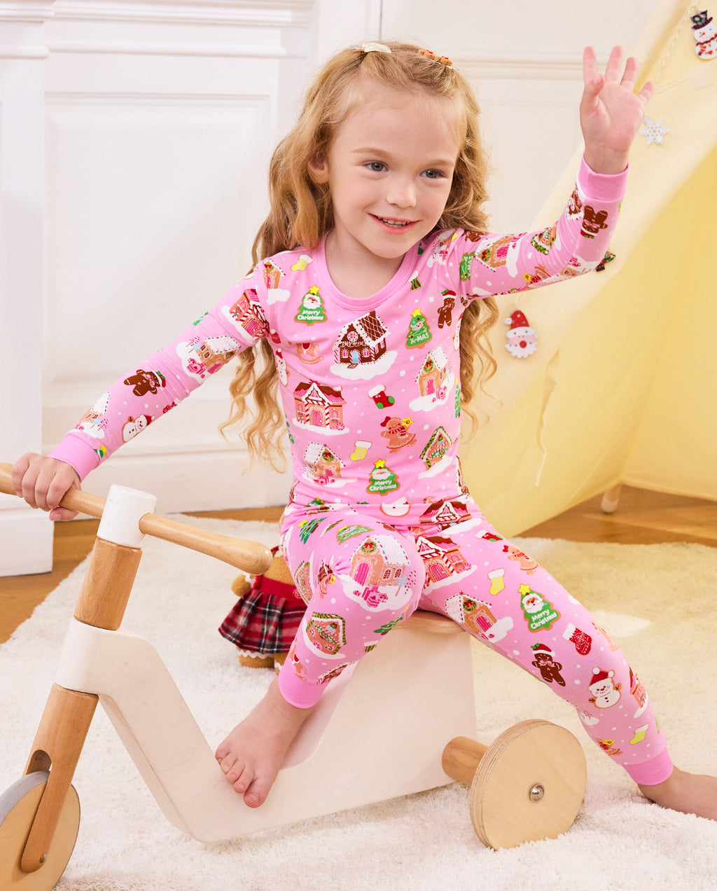 Child raising one hand while riding a wooden tricycle in Gingerbread Lane pajamas.