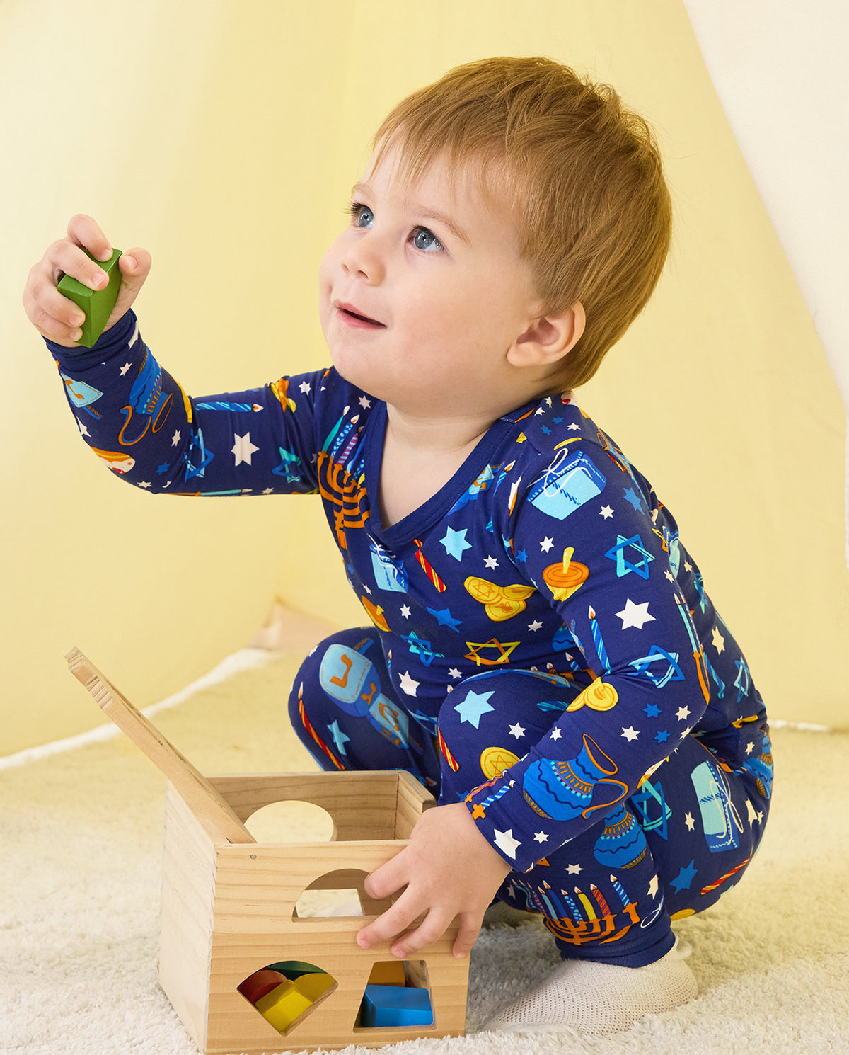 Child holding a green toy block beside a wooden tray in the Hanukkah Magic set.