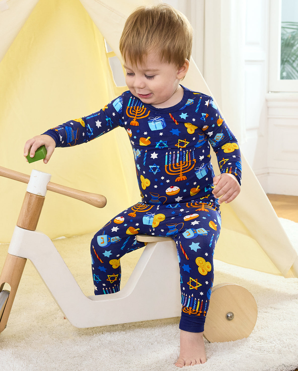 Child seated on a wooden tricycle holding a toy block in the Hanukkah Magic pajama set.