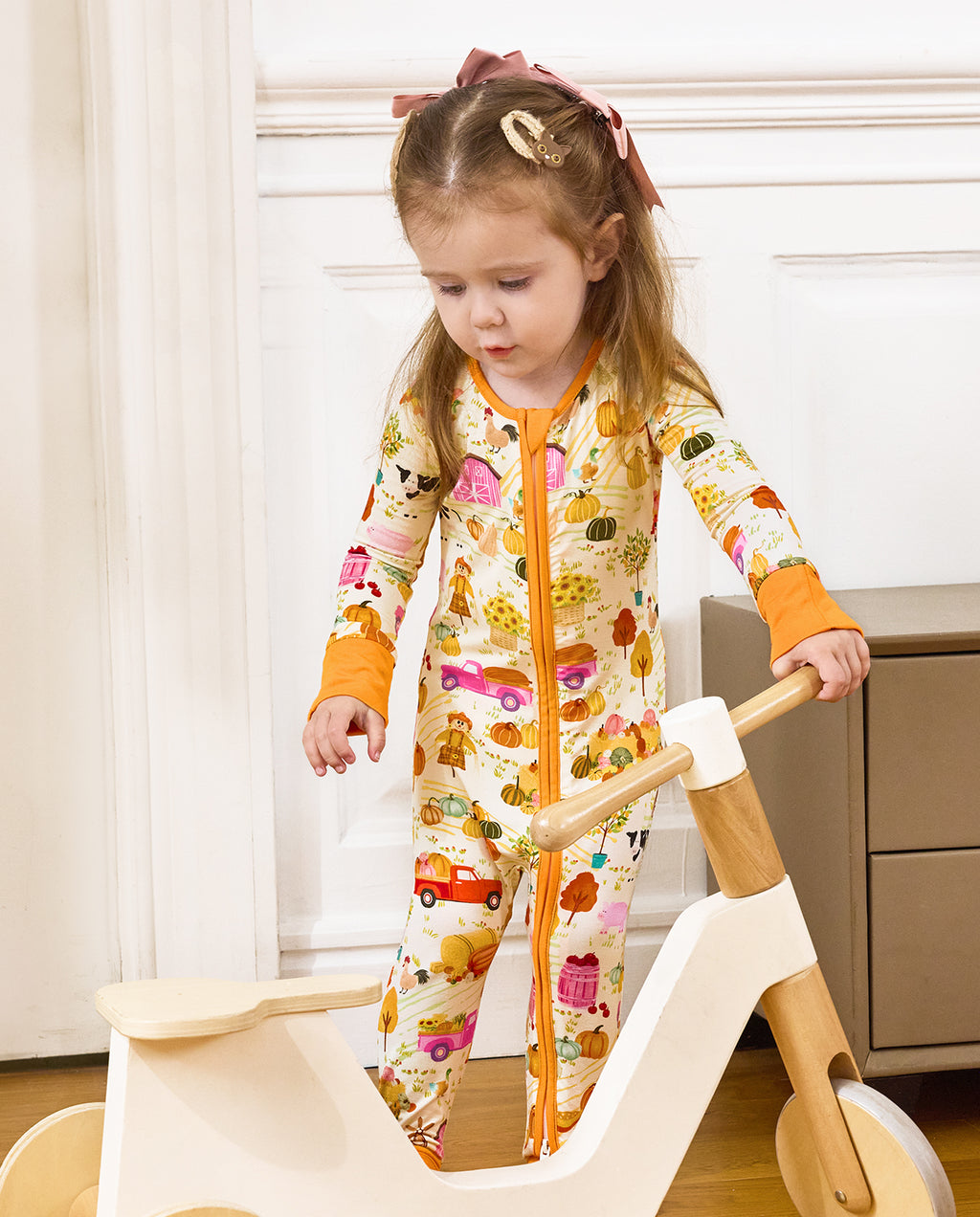 Front view of a child standing beside a wooden rocker in the Harvest Farm romper.