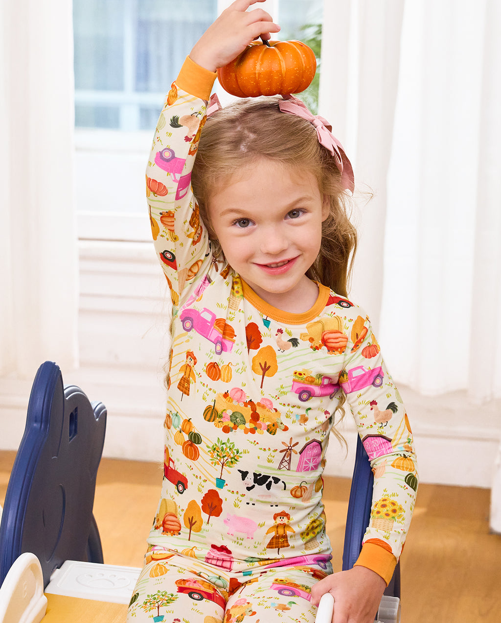 Child holding a pumpkin overhead in cream Harvest Farm pajamas with orange trim.