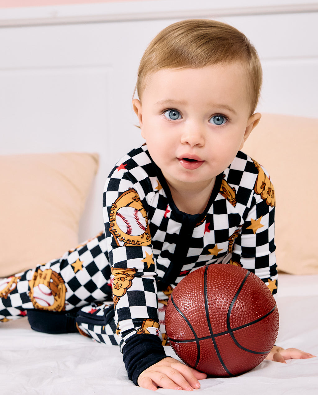 A baby boy wearing a black and white checkered pajama set with baseball and glove graphics, lying on a white bed and holding a small basketball.