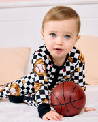 A baby boy wearing a black and white checkered pajama set with baseball and glove graphics, lying on a white bed and holding a small basketball.