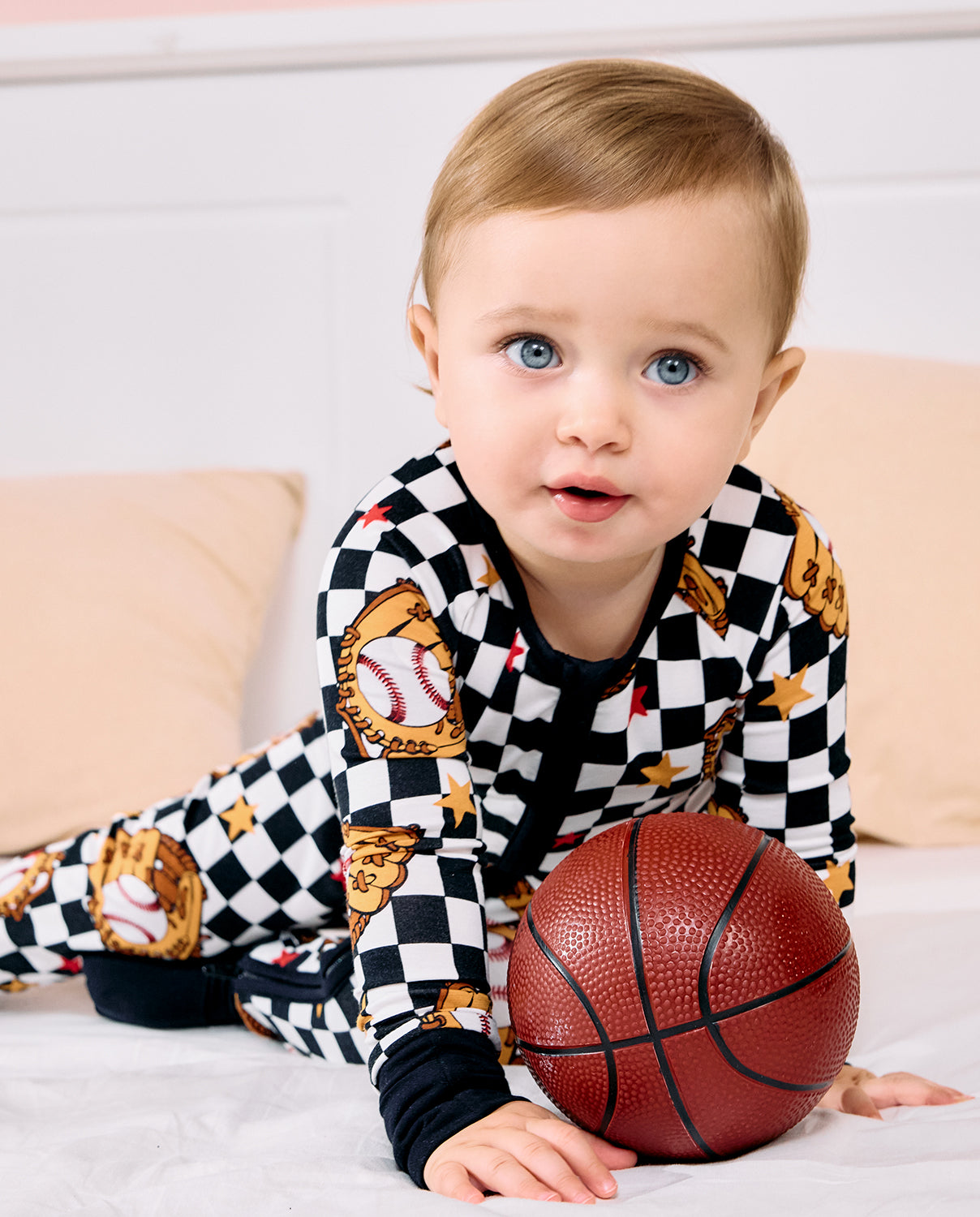 A baby boy wearing a black and white checkered pajama set with baseball and glove graphics, lying on a white bed and holding a small basketball.