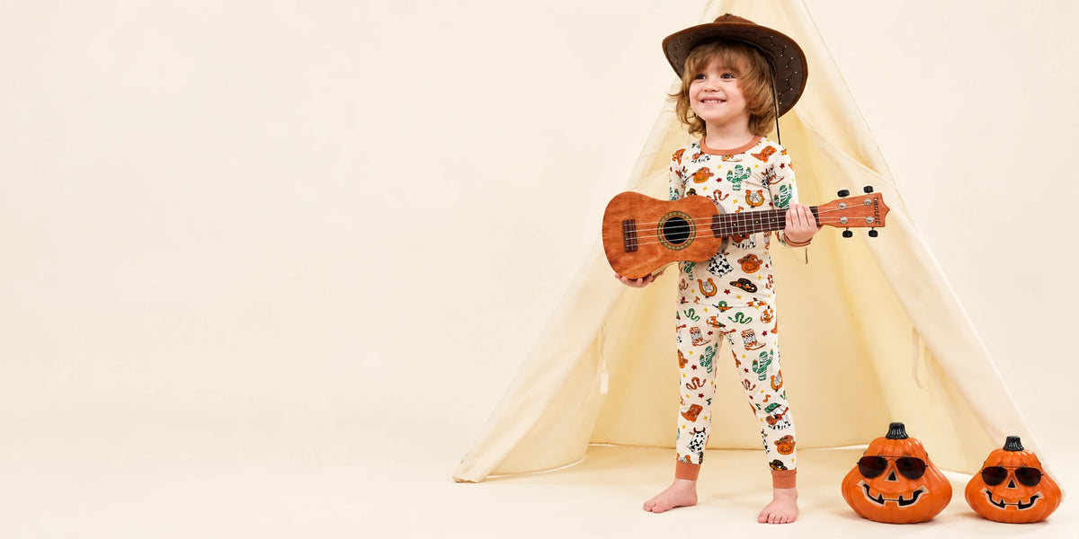 Young child wearing black Halloween zippy pajamas in a warm home scene with pumpkins, bats, and glowing decorations.