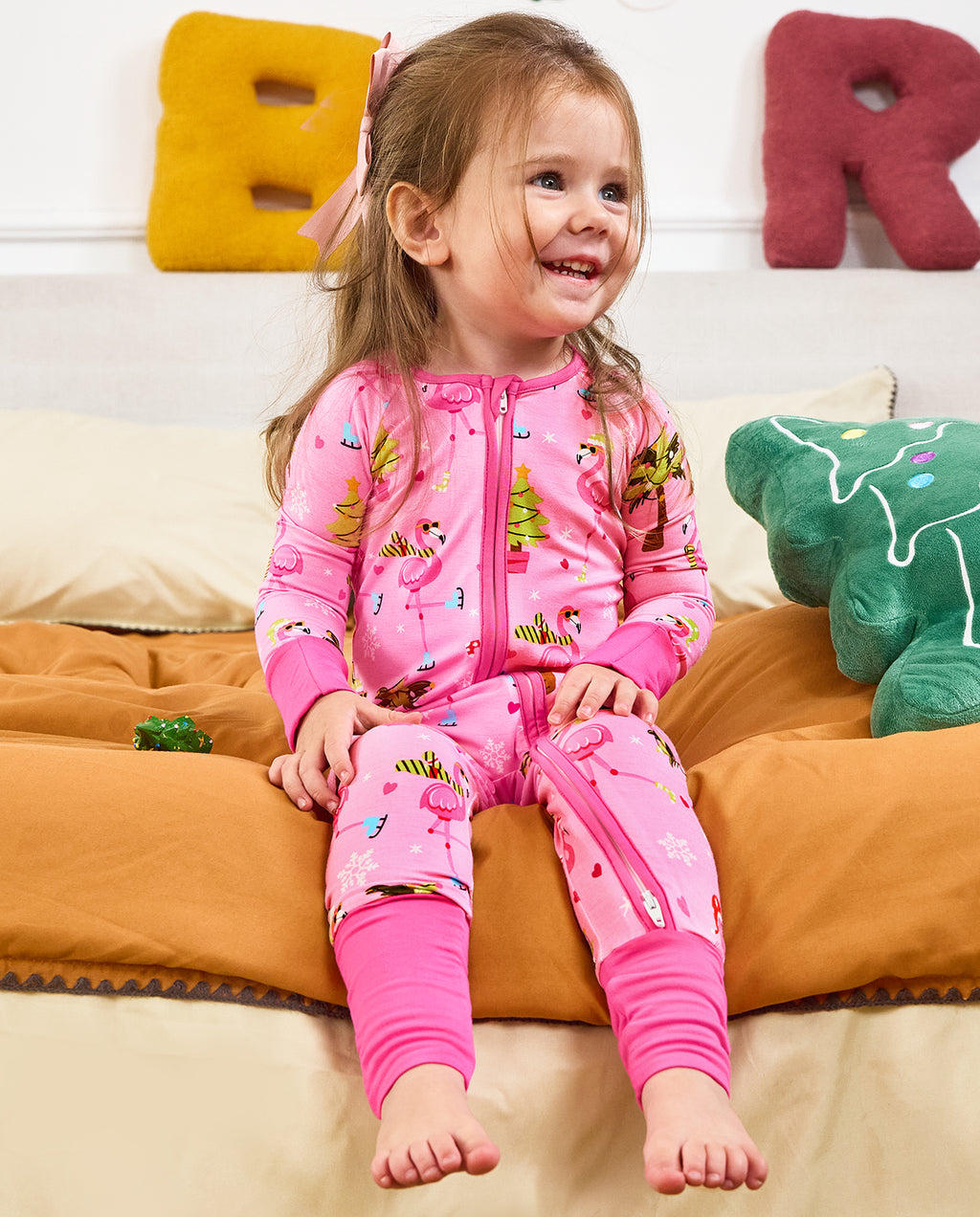 Child seated on orange bedding smiling in the Merry Mingo romper with uncovered feet.