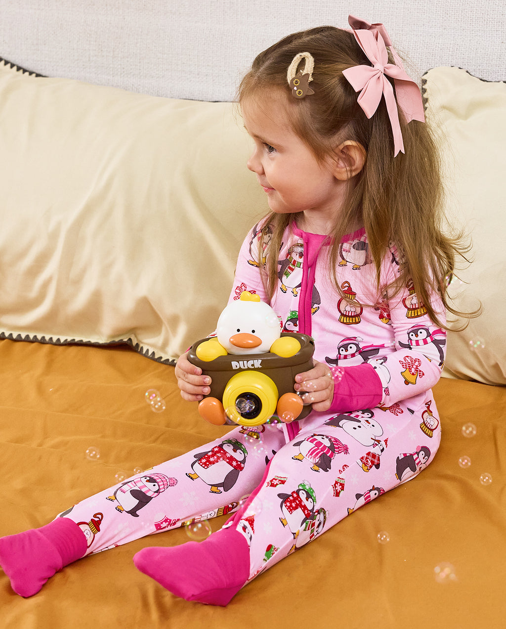 Child seated on bedding with a toy camera in the Merry Penguins romper.
