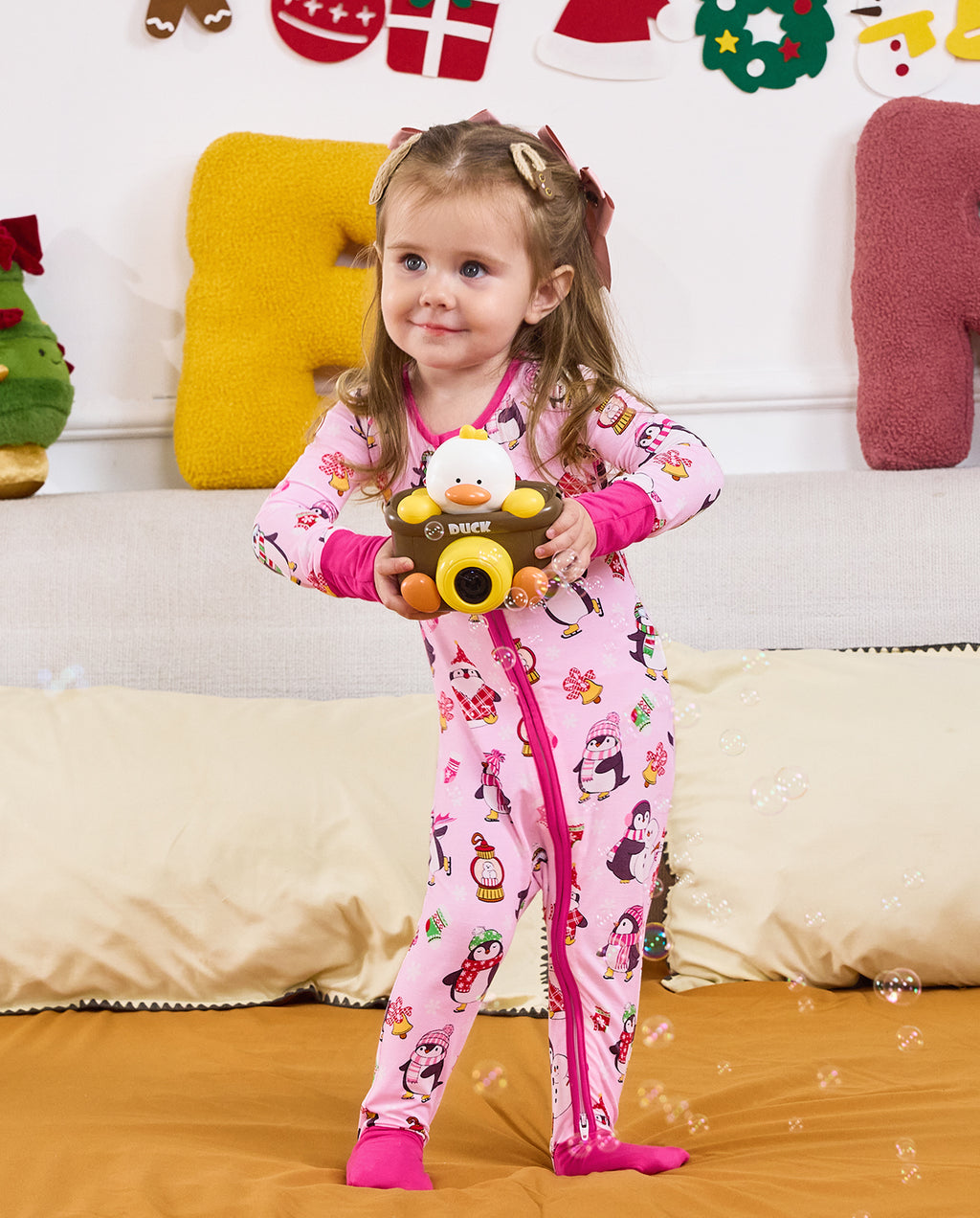 Child standing by a sofa holding a toy camera in the Merry Penguins romper.