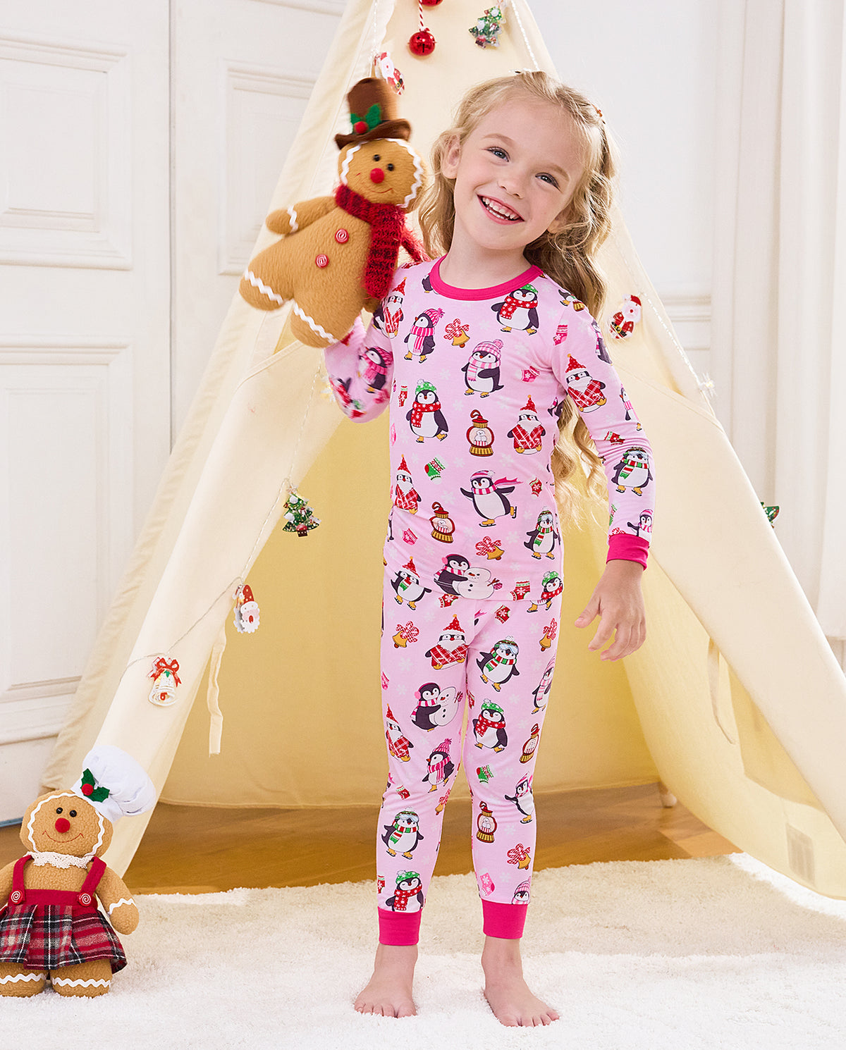 Child standing by a holiday play tent with a gingerbread plush in Merry Penguins pajamas.