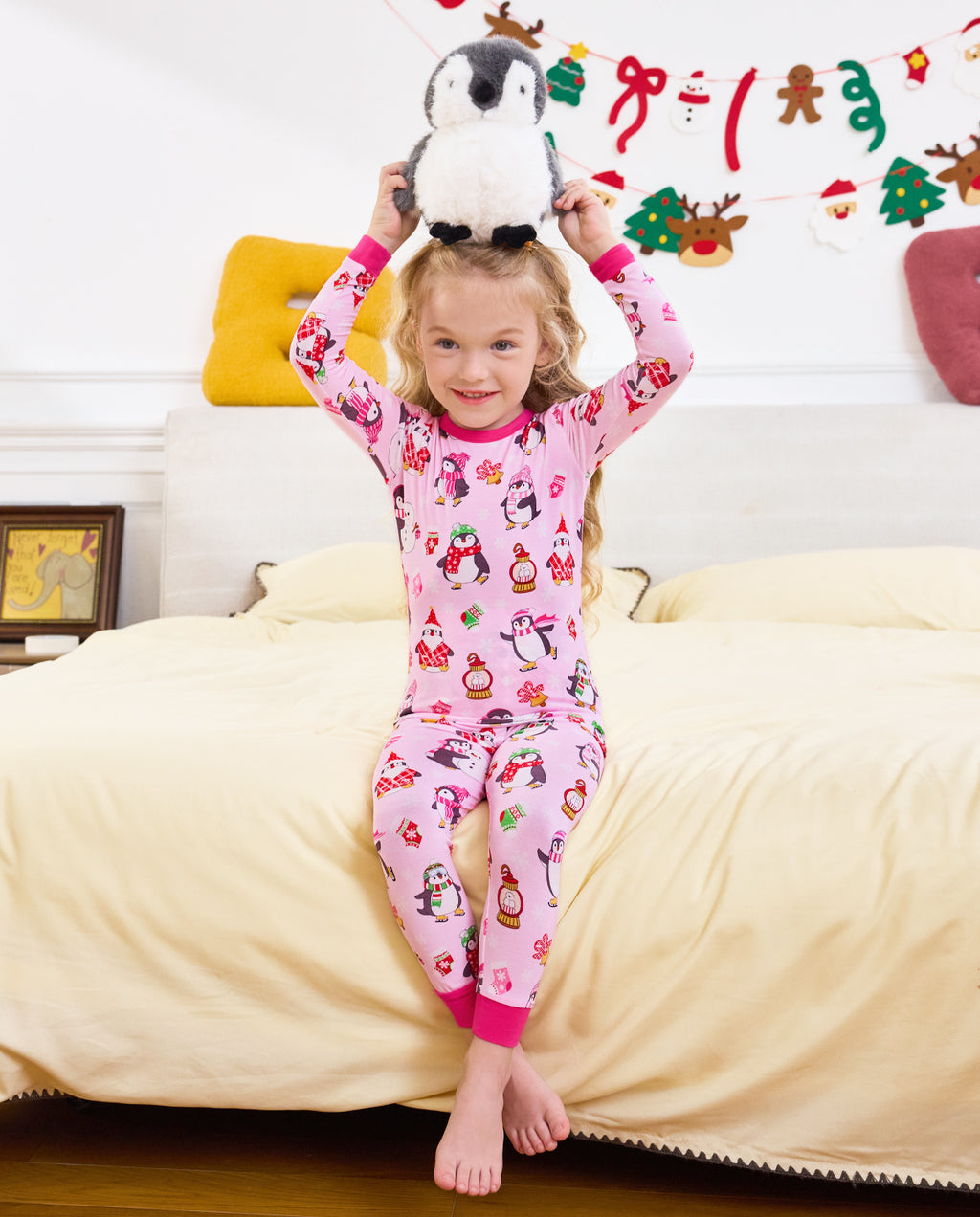 Full view of a child standing on bedding with a penguin plush overhead in Merry Penguins pajamas.
