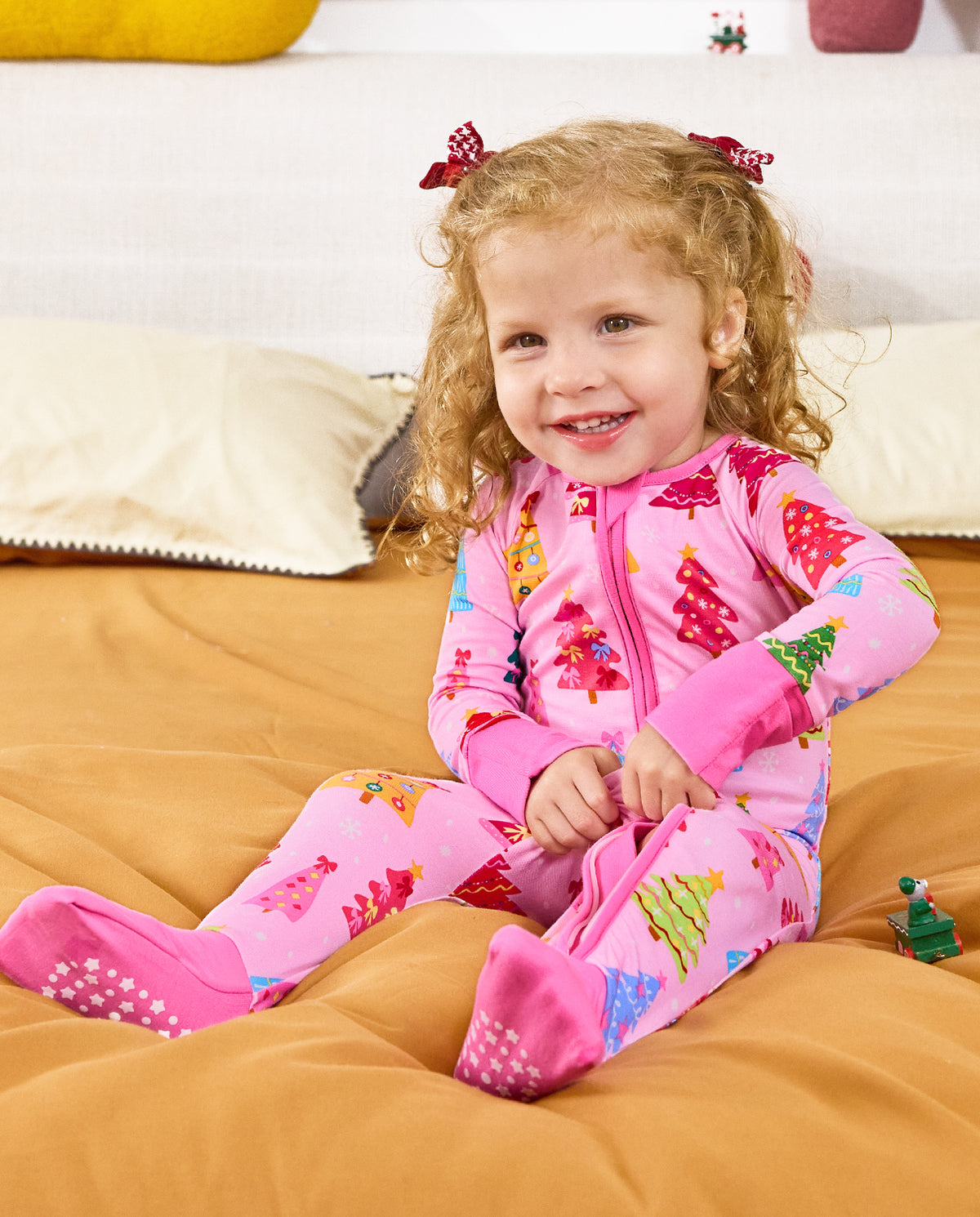 Child seated on bedding and smiling in the pink Pastel Christmas romper.
