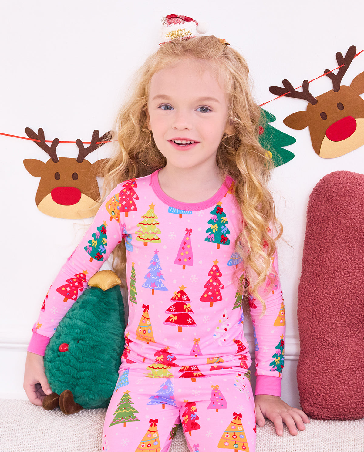 Smiling child seated on a sofa holding a plush tree in Pastel Christmas pajamas.