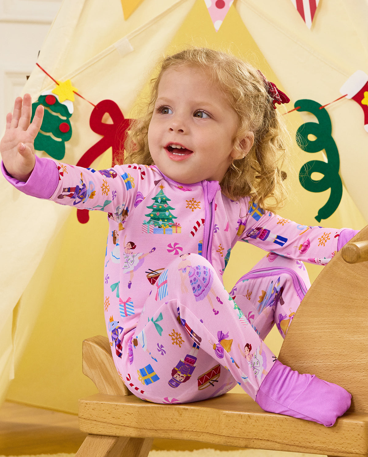 Child seated and waving with a toy in the Pink Nutcracker romper.