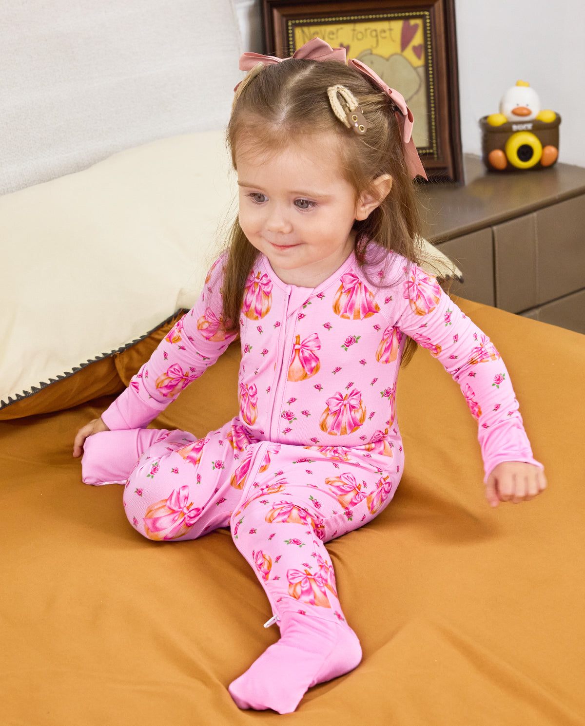 Child seated on orange bedding in the pink Pumpkin Bows zip-up romper with covered feet.