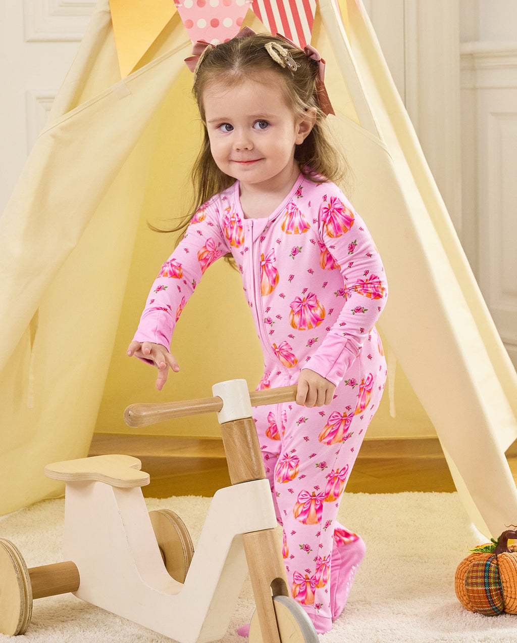 Child standing by a wooden tricycle beside a play tent in the Pumpkin Bows romper.