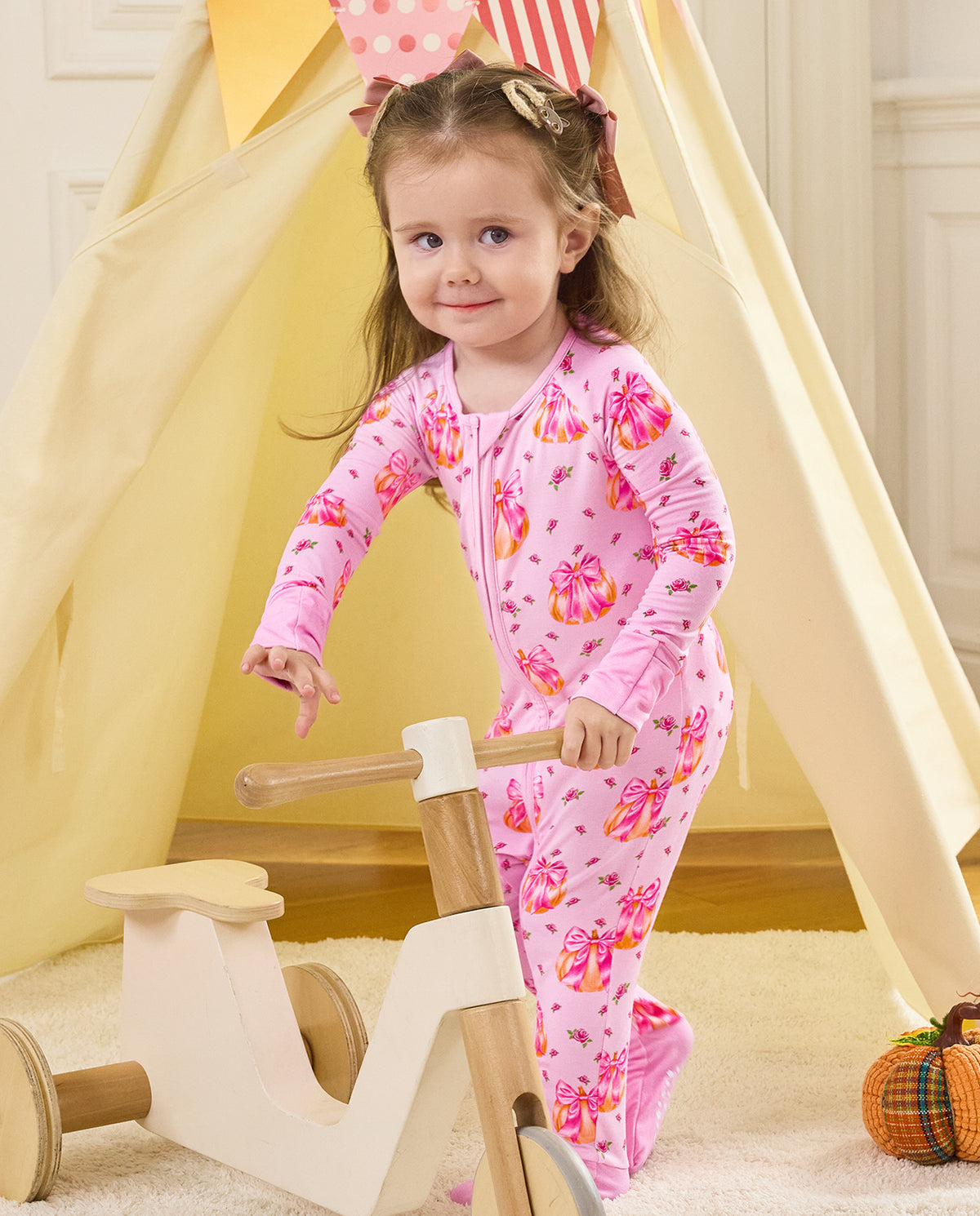 Child standing by a wooden tricycle beside a play tent in the Pumpkin Bows romper.