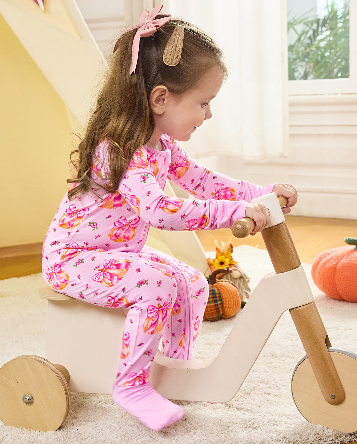 Side view of a child seated on a wooden tricycle wearing the Pumpkin Bows romper.