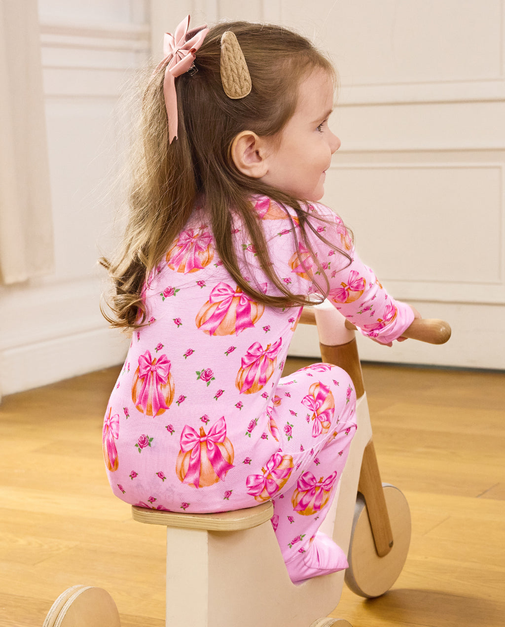 Rear view of a child seated on a wooden tricycle in the pink Pumpkin Bows romper.
