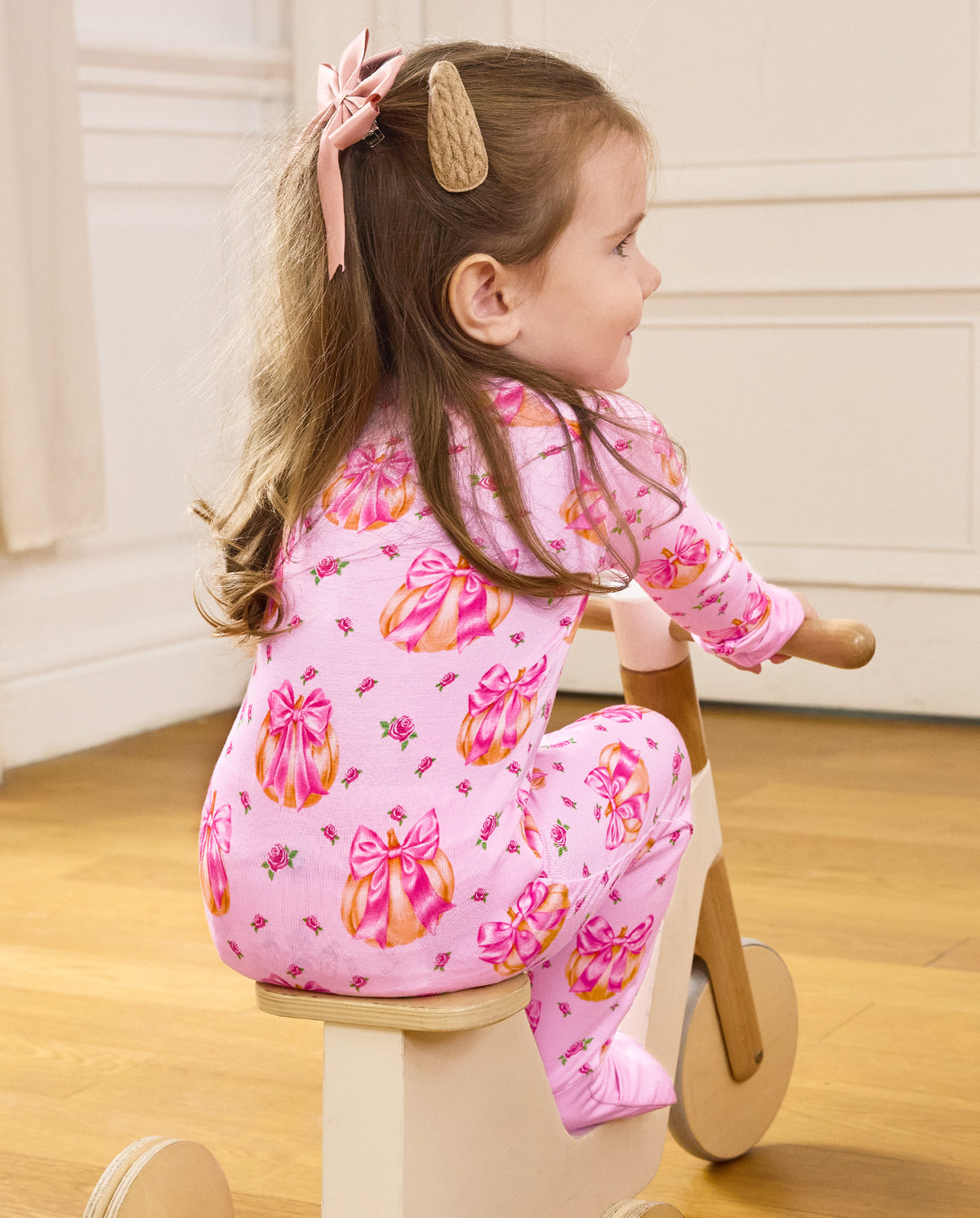 Rear view of a child seated on a wooden tricycle in the pink Pumpkin Bows romper.