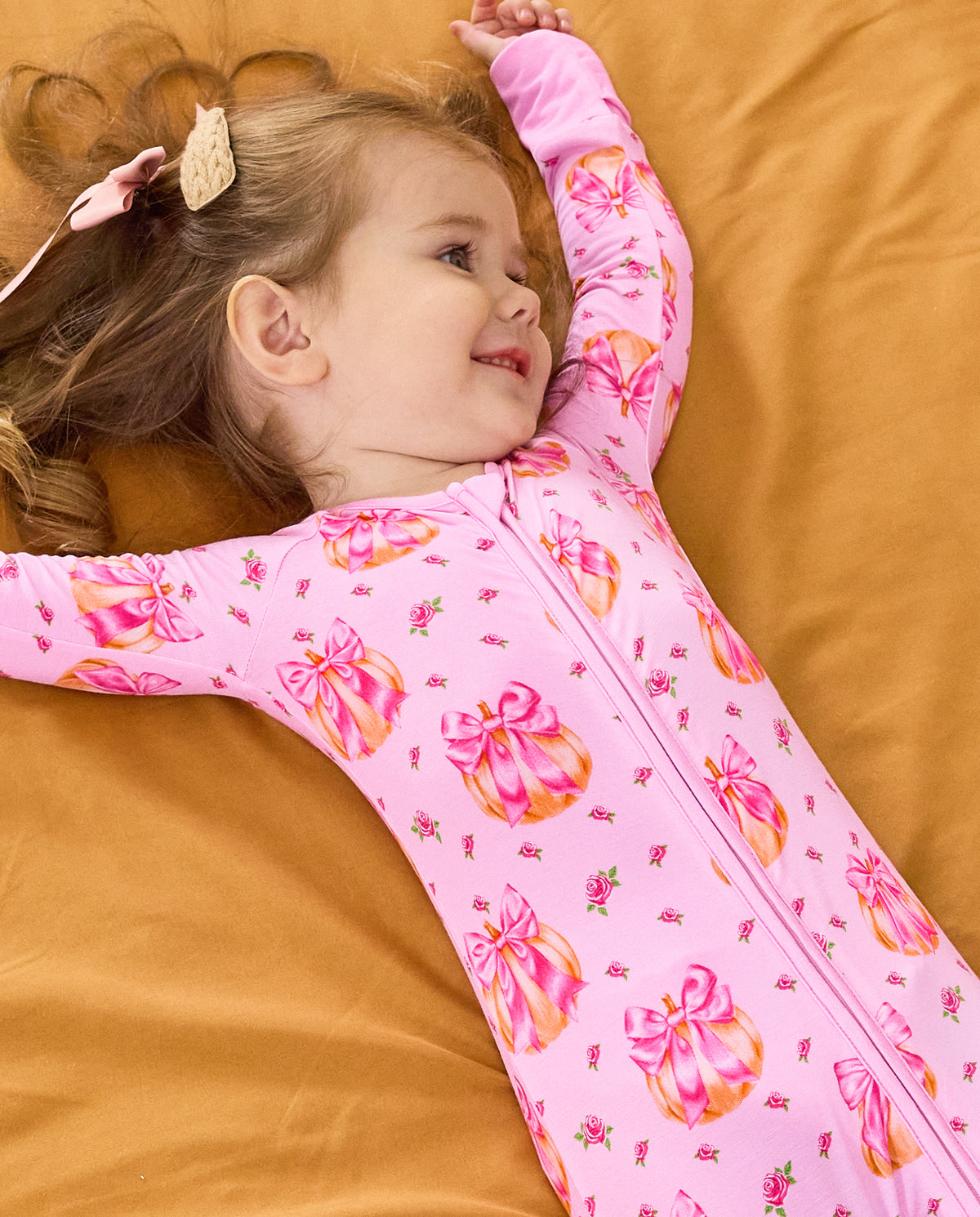 Child lying on orange bedding with arms raised in the Pumpkin Bows romper.
