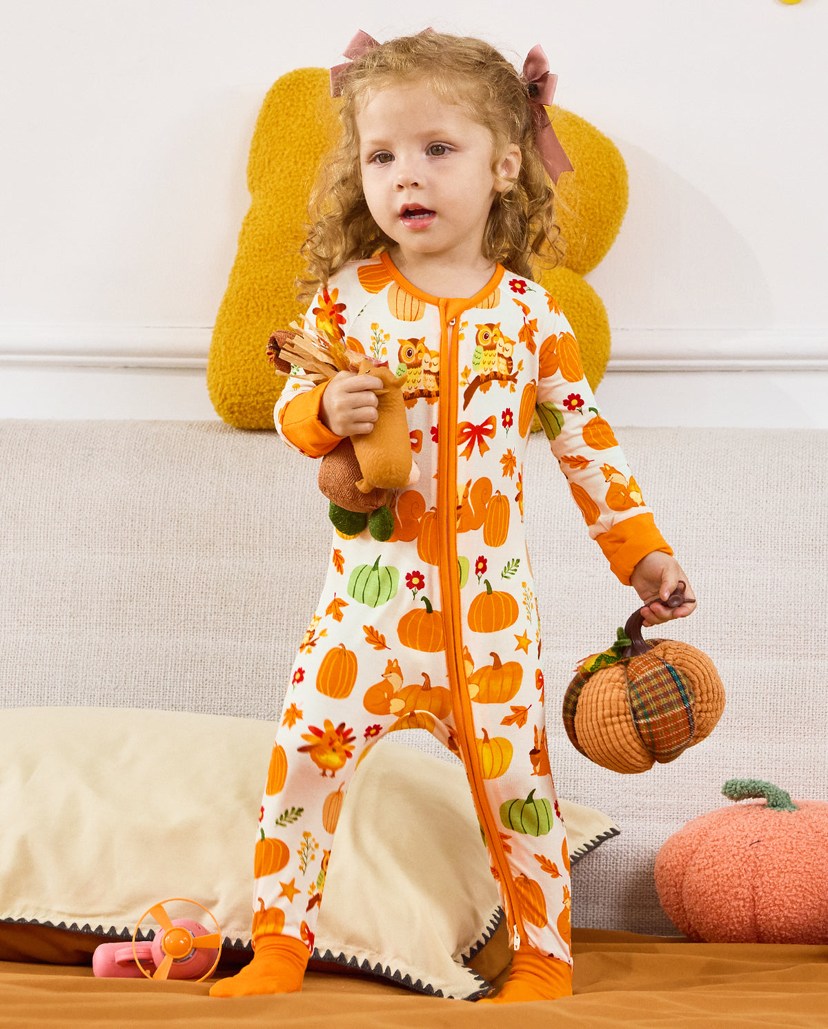Child standing with a pinecone toy and a pumpkin decoration in the Pumpkin Patch romper.
