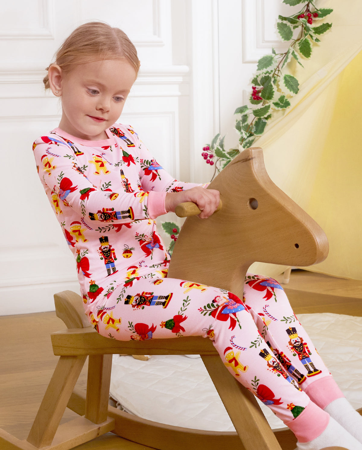 Child looking downward while seated on a wooden rocking horse in Sugarplum Wishes pajamas.