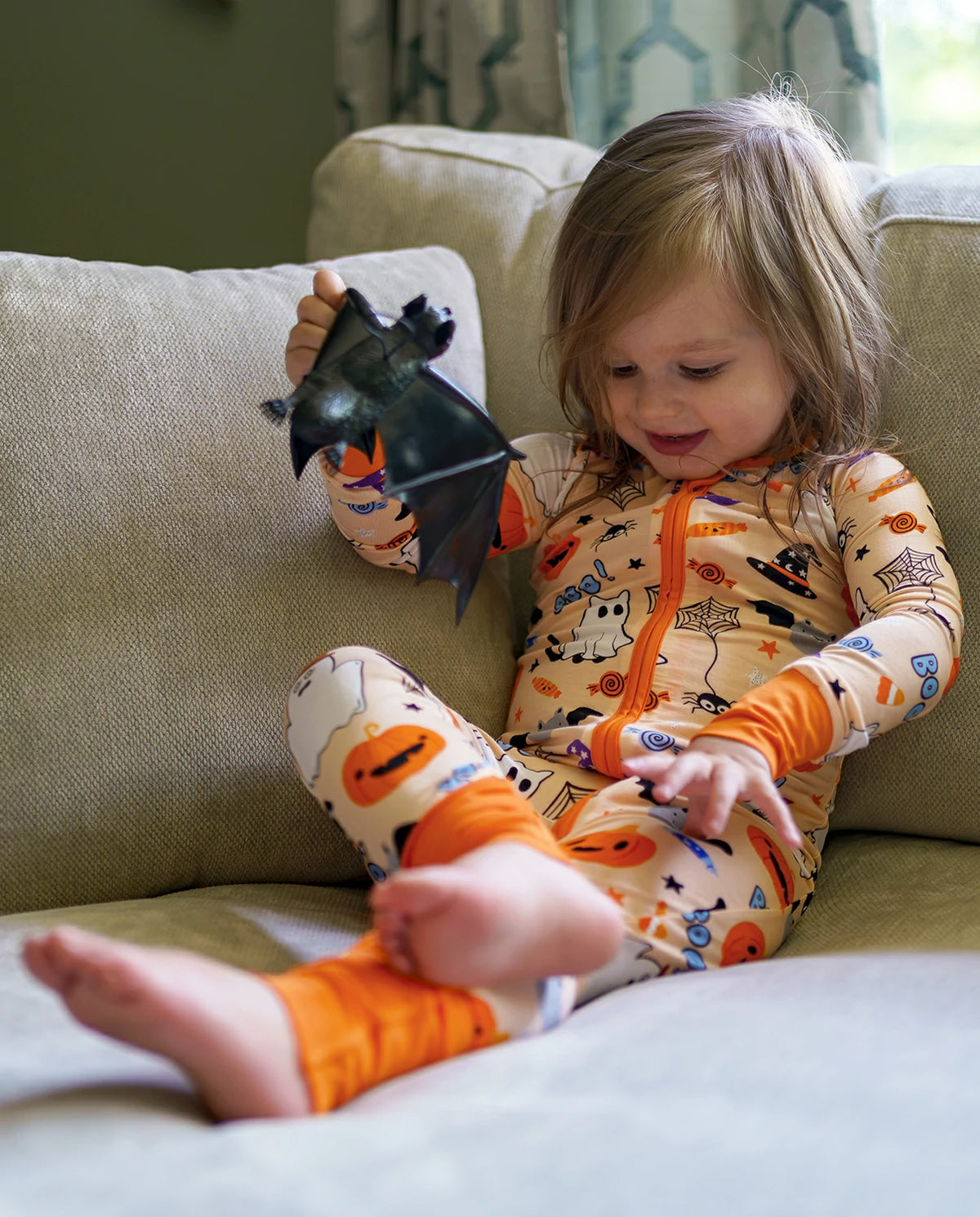 Child playing with a black bat toy while wearing Boo Treats Bamboo Zippy on a couch