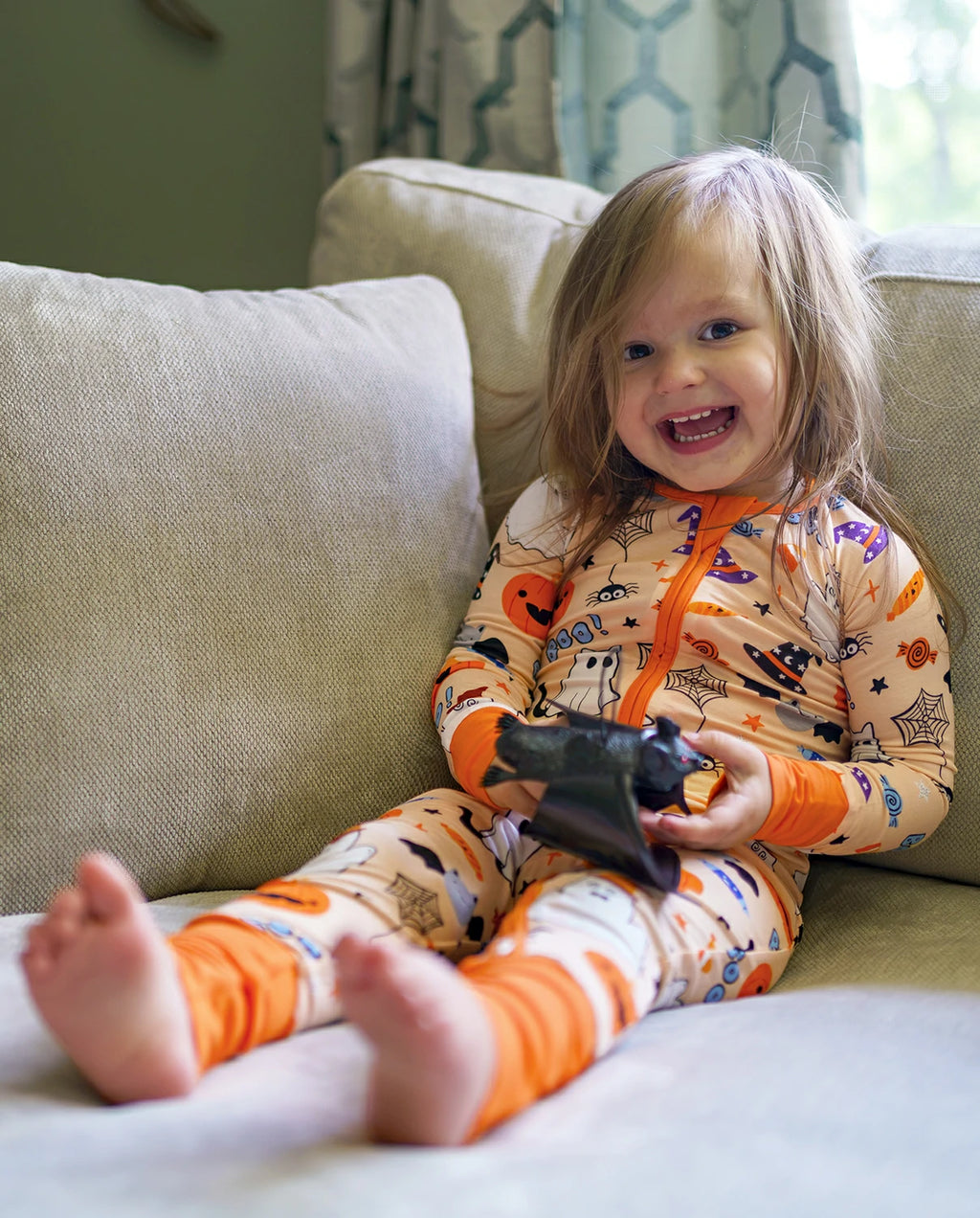 Child smiling on couch in Boo Treats Bamboo Zippy with bat toy and Halloween print