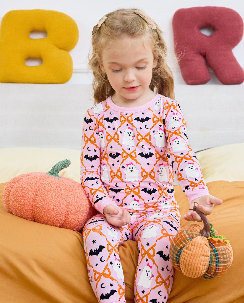 Ghost Bows Long Two-Piece Pajama Set - child wearing the ghost-and-bow pajamas while seated with plush pumpkins