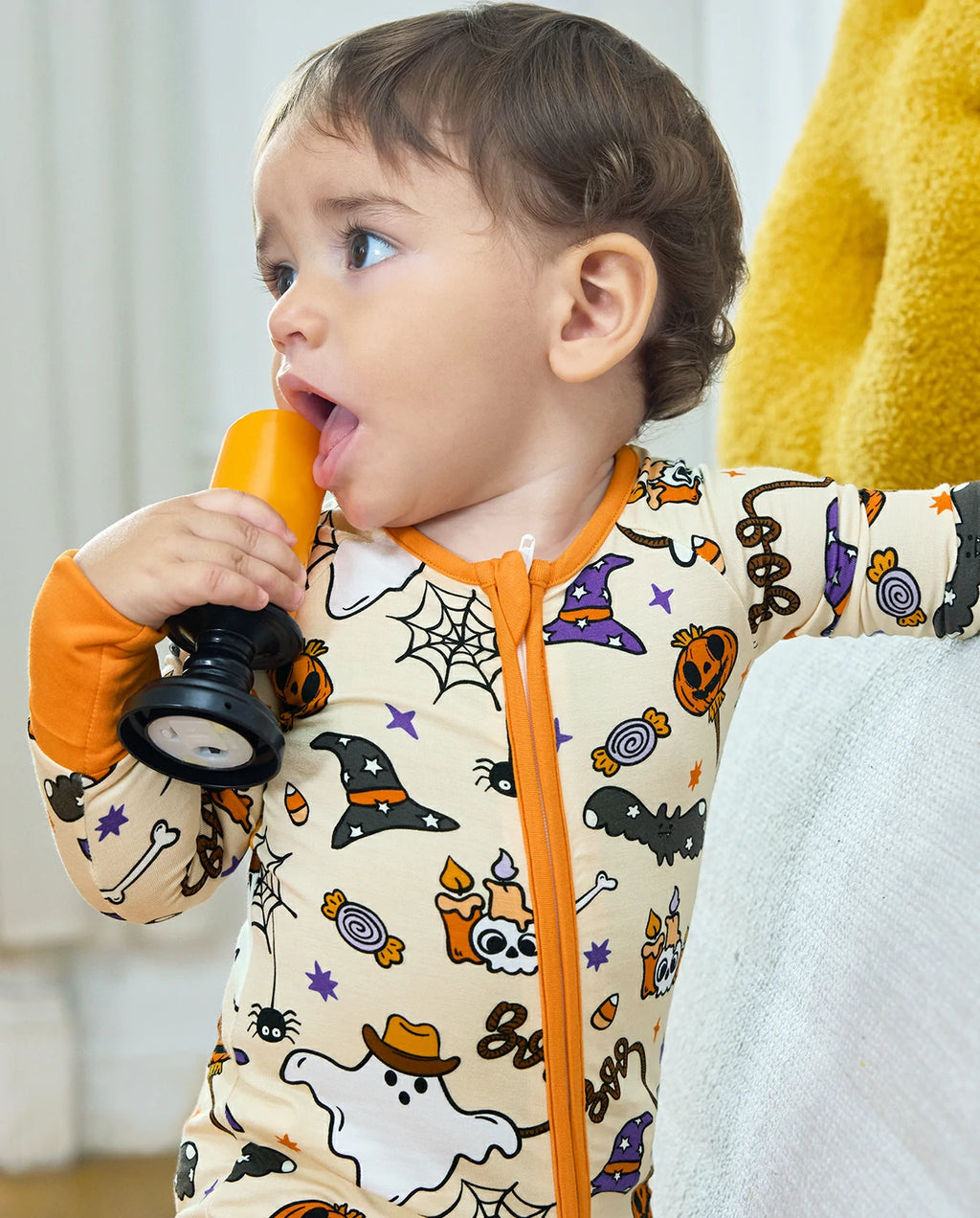A baby wearing a cream-colored Halloween-themed zip-up sleeper with orange trim and a spiderweb print, holding a toy flashlight.