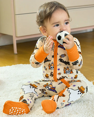 A baby sitting on a white rug wearing a beige and orange Halloween-themed footed onesie, holding a small soccer ball to their mouth.