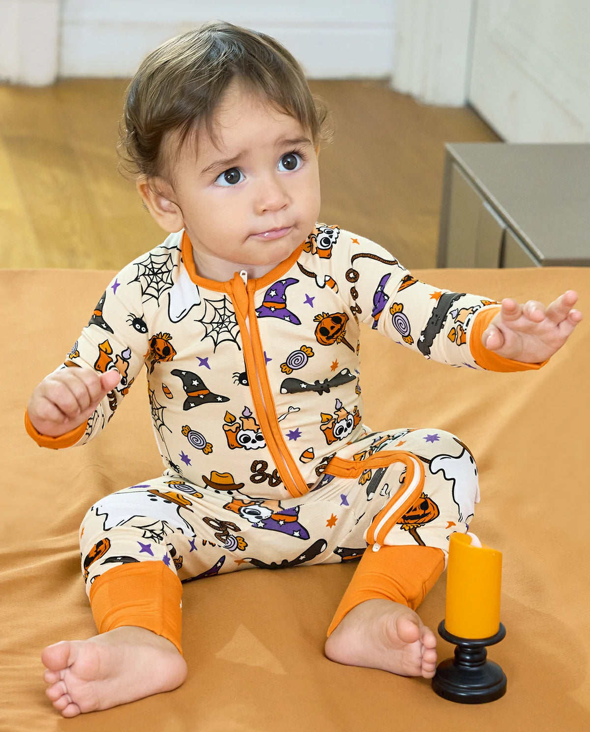 A baby sitting on an orange blanket wearing a Halloween-themed cream-colored one-piece pajama with orange trim and a full-length zipper. The pajama features a print of witches, bats, pumpkins, and spiderwebs.