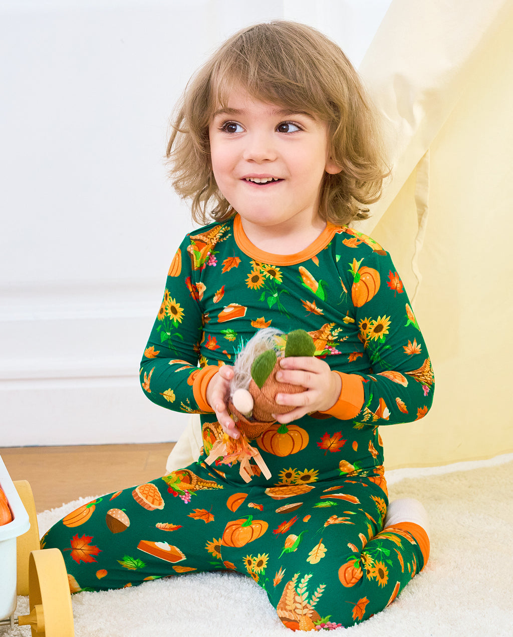 Child seated in Harvest Feast long two-piece pajamas with orange trim while holding a plush harvest toy.