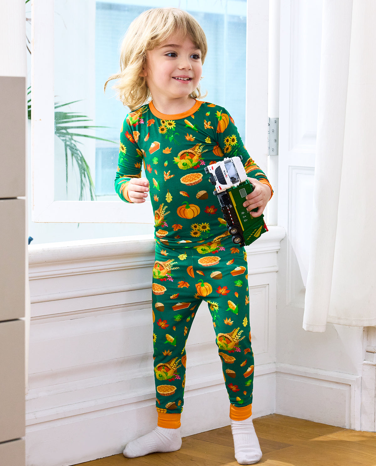 Child wearing Harvest Feast long two-piece pajamas and holding a green toy truck.