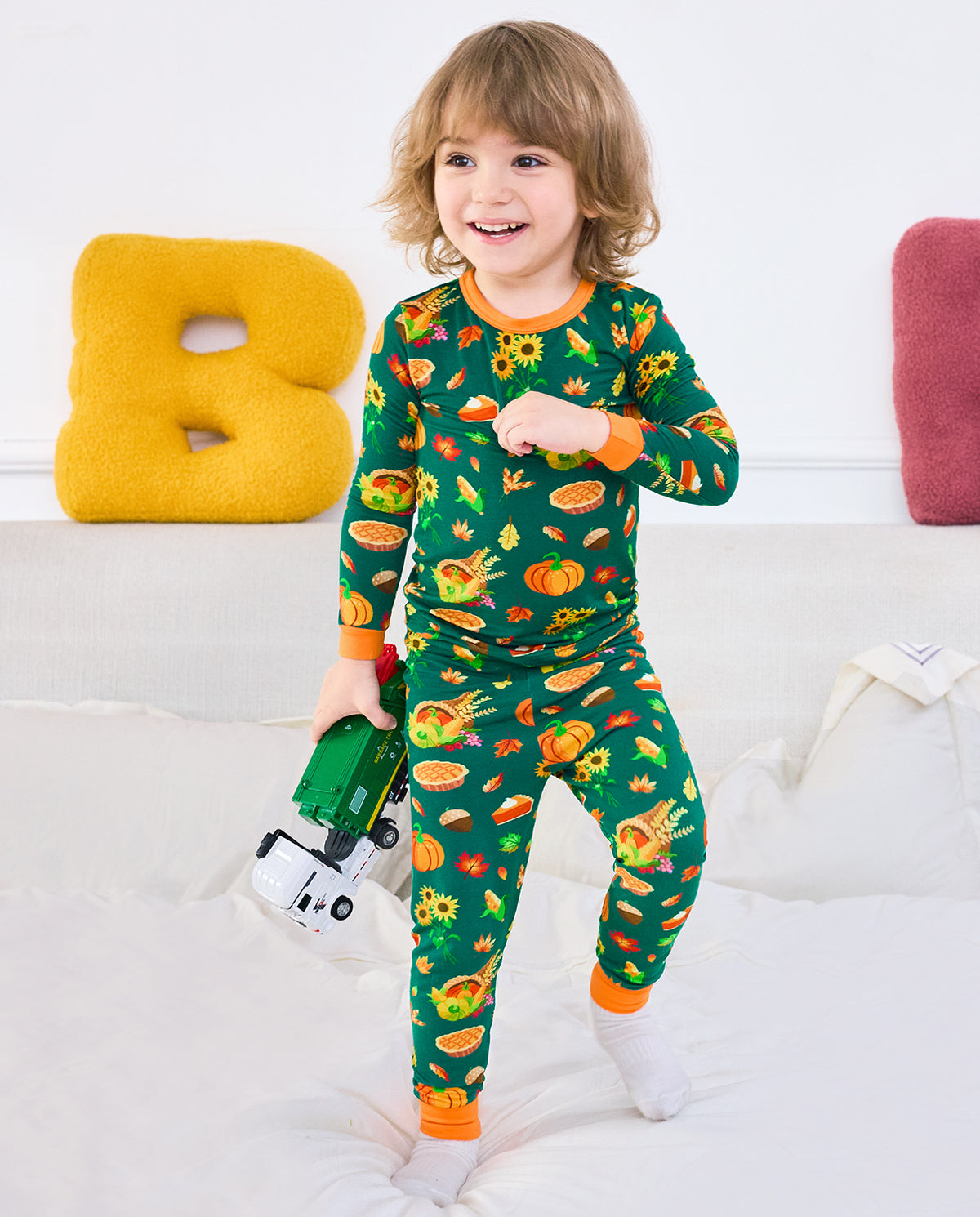 Child standing on a bed in Harvest Feast long two-piece pajamas while holding toy trucks.